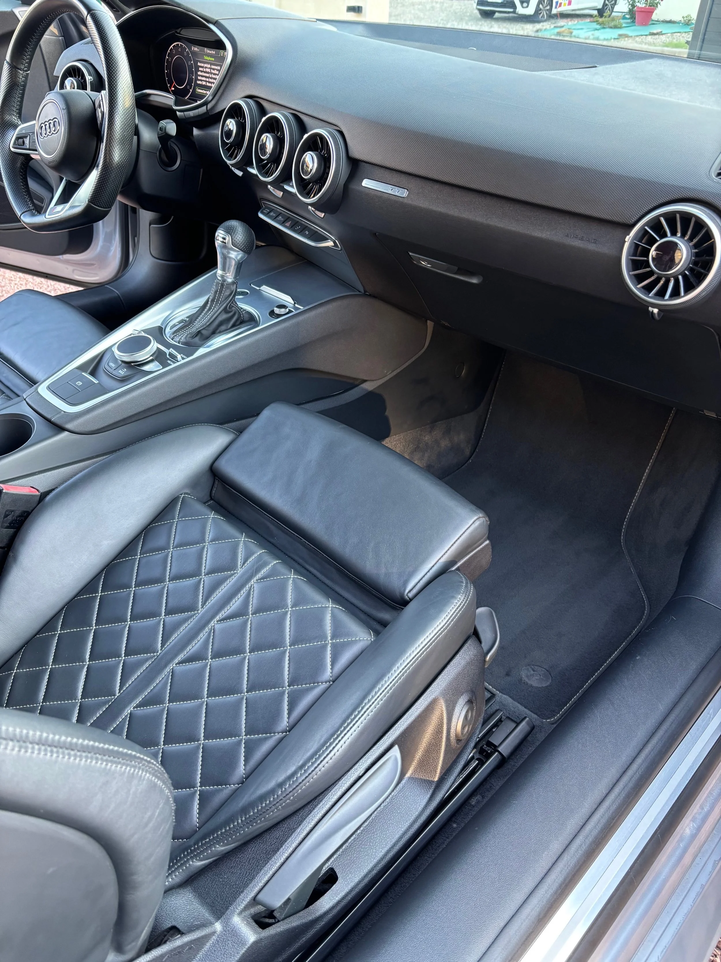 Intérieur d'une voiture Audi, avec tableau de bord, volant, volant, console centrale avec levier de vitesse, siège en cuir noir avec motif de couture en diamant, et un côté du véhicule visible.