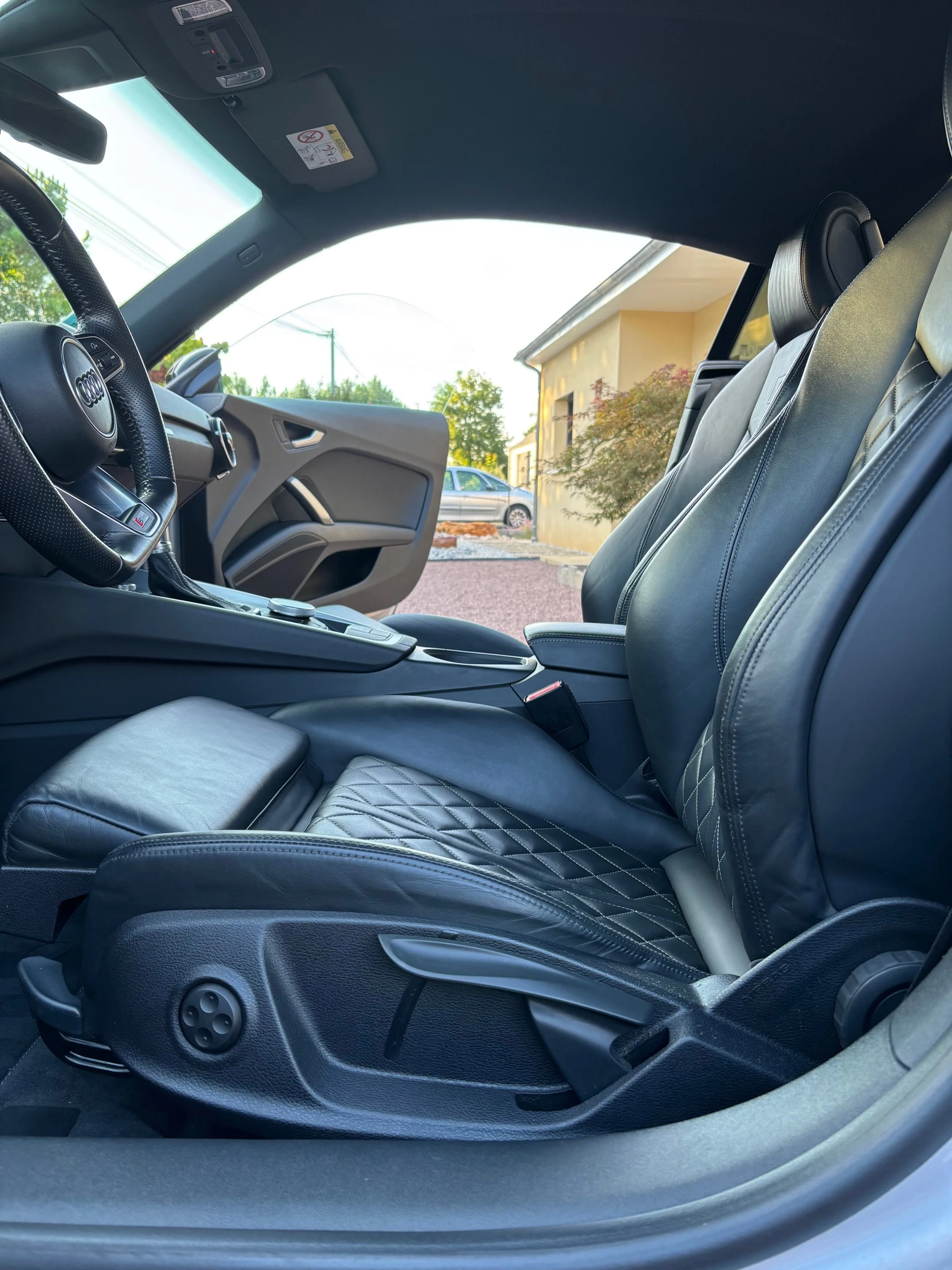 Intérieur d'une voiture de luxe, mettant en valeur les sièges en cuir noir avec motifs en losange, tableau de bord moderne et porte ouverte avec vue sur un bâtiment résidentiel et une voiture à l'extérieur.