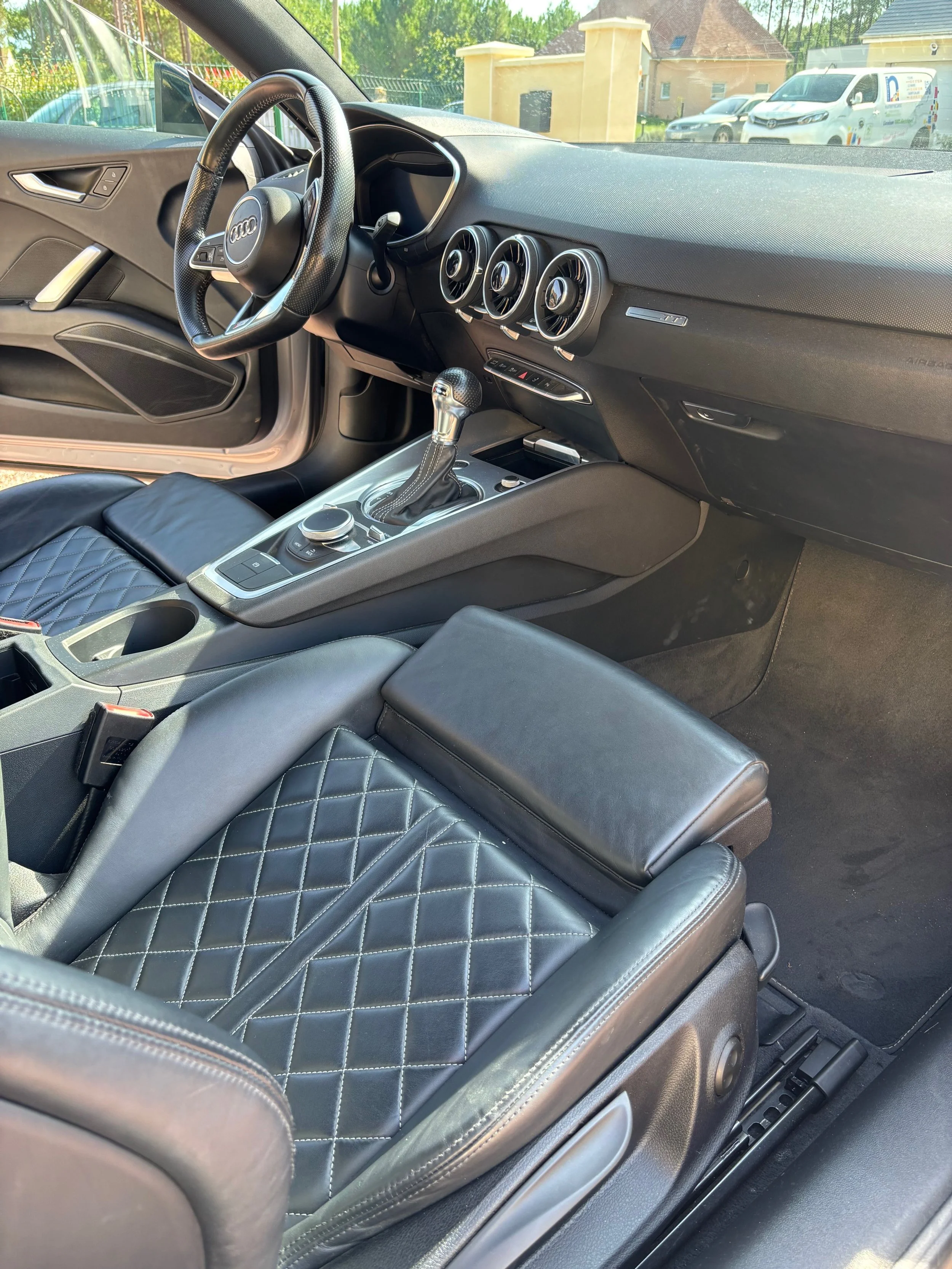 Intérieur d'une voiture noire avec des sièges en cuir noir à motif de couture en diamant. Le tableau de bord est équipé de trois bouches d'aération circulaires. Le levier de vitesses est en position manuelle. Il y a un bouton de contrôle sur la console centrale, ainsi qu'un cadran de commande. La voiture est stationnée à l'extérieur, avec une vue de maisons et d'autres véhicules par la fenêtre.