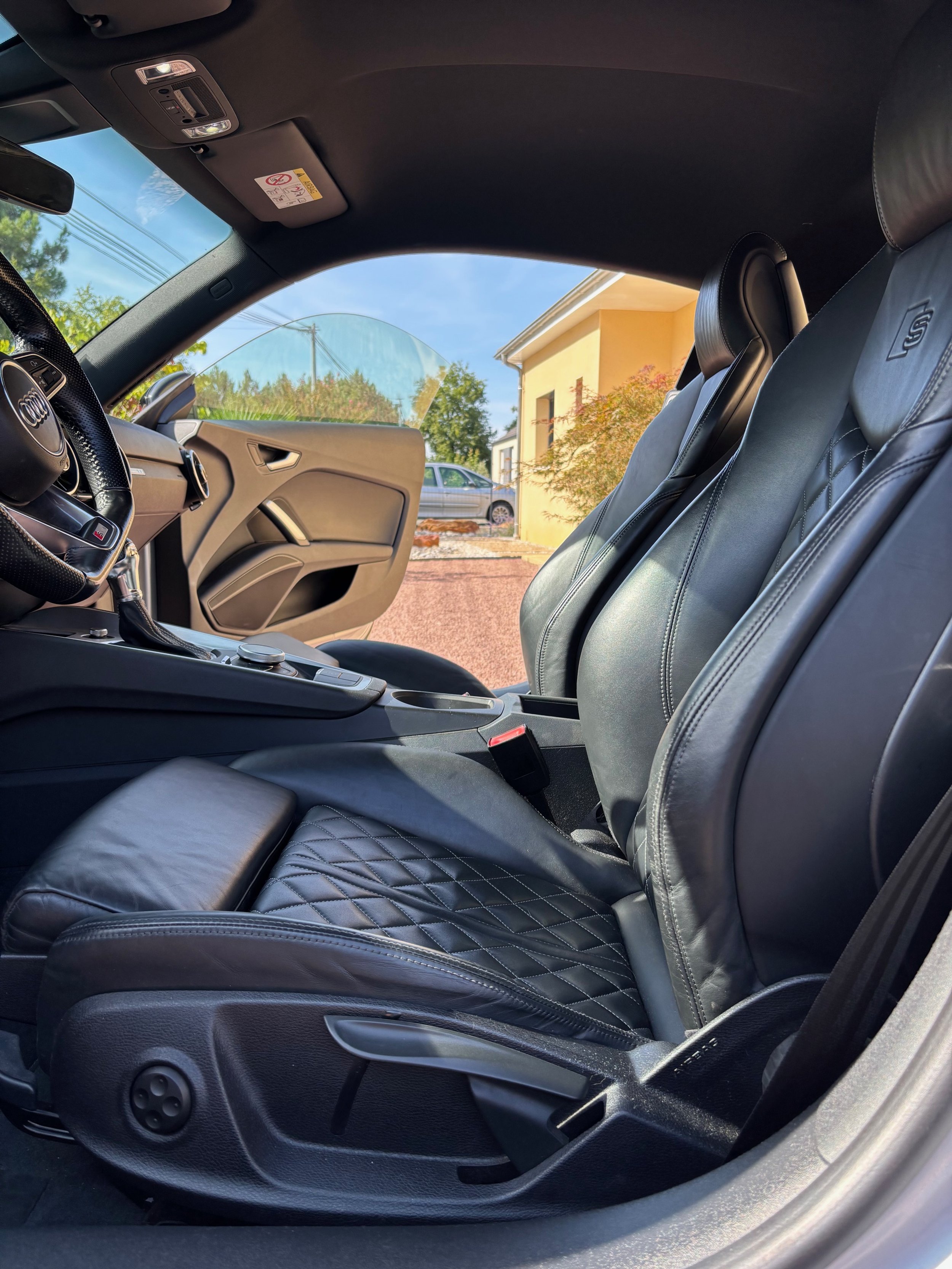 Intérieur d'une voiture sportive avec sièges en cuir noir, tableau de bord visible, porte côté conducteur ouverte, extérieure visible avec un bâtiment, un arbre et une autre voiture en arrière-plan.