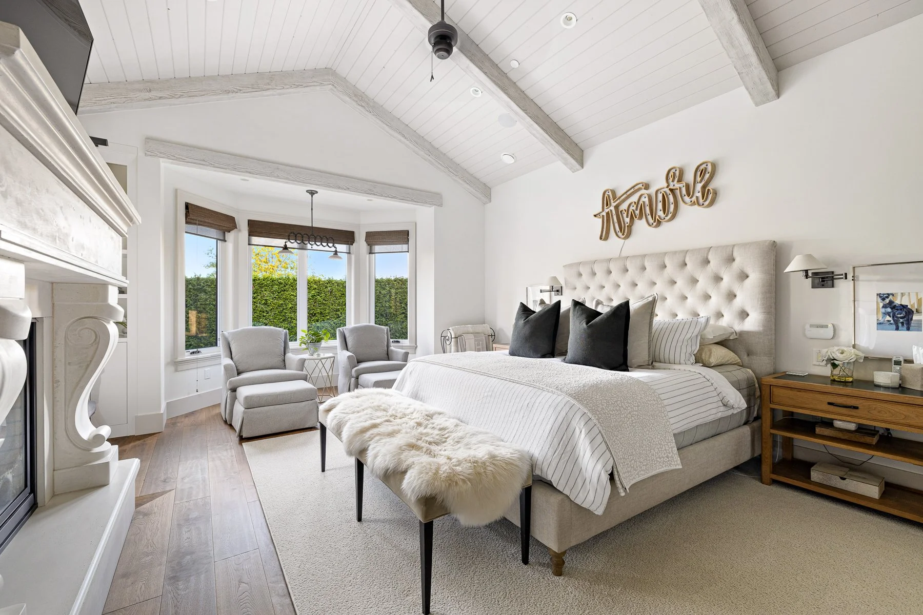 Bright bedroom with a large upholstered bed, decorative pillows, and a headboard wall art that reads "amour." There are two wall-mounted lamps on either side of the bed, a wooden nightstand with a plant and books, and a fluffy bench at the foot of the bed. The room features a vaulted ceiling with exposed beams, a seating area with two chairs and a small table near large bay windows, and light-colored walls and flooring.