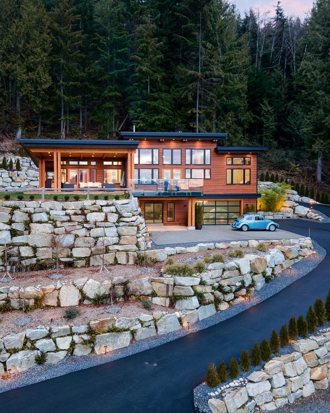 Modern multi-story house with large windows, wooden exterior, and a balcony, situated on a sloped lot with stone retaining walls, a driveway with a light blue vintage Volkswagen Beetle, surrounded by tall trees.