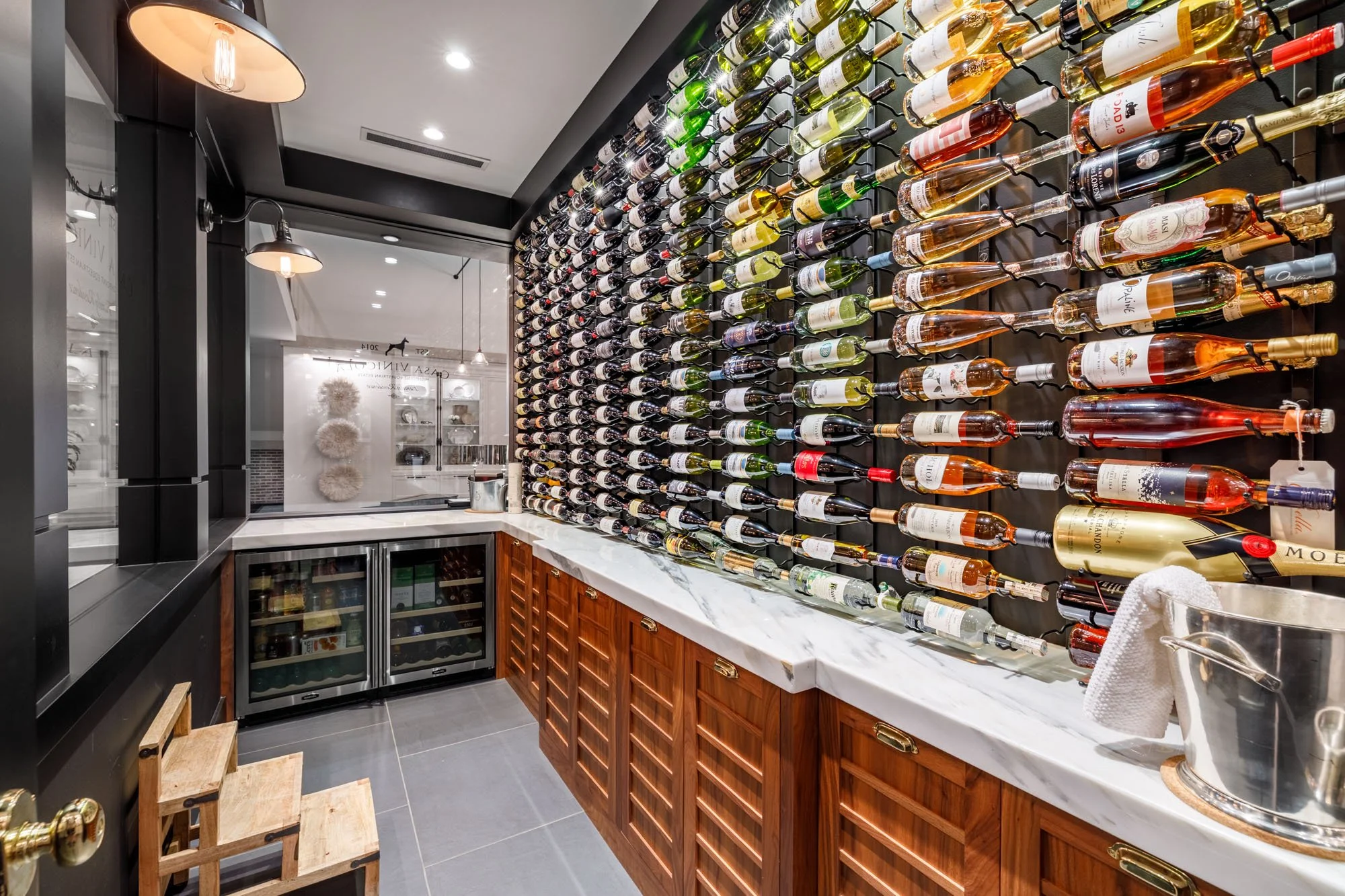 A wine storage area with a wall-mounted wine rack displaying numerous bottles of wine, a marble countertop, and a small wine refrigerator beneath. There are wooden chairs on the floor near the counter, and a bucket with a towel on the right side.
