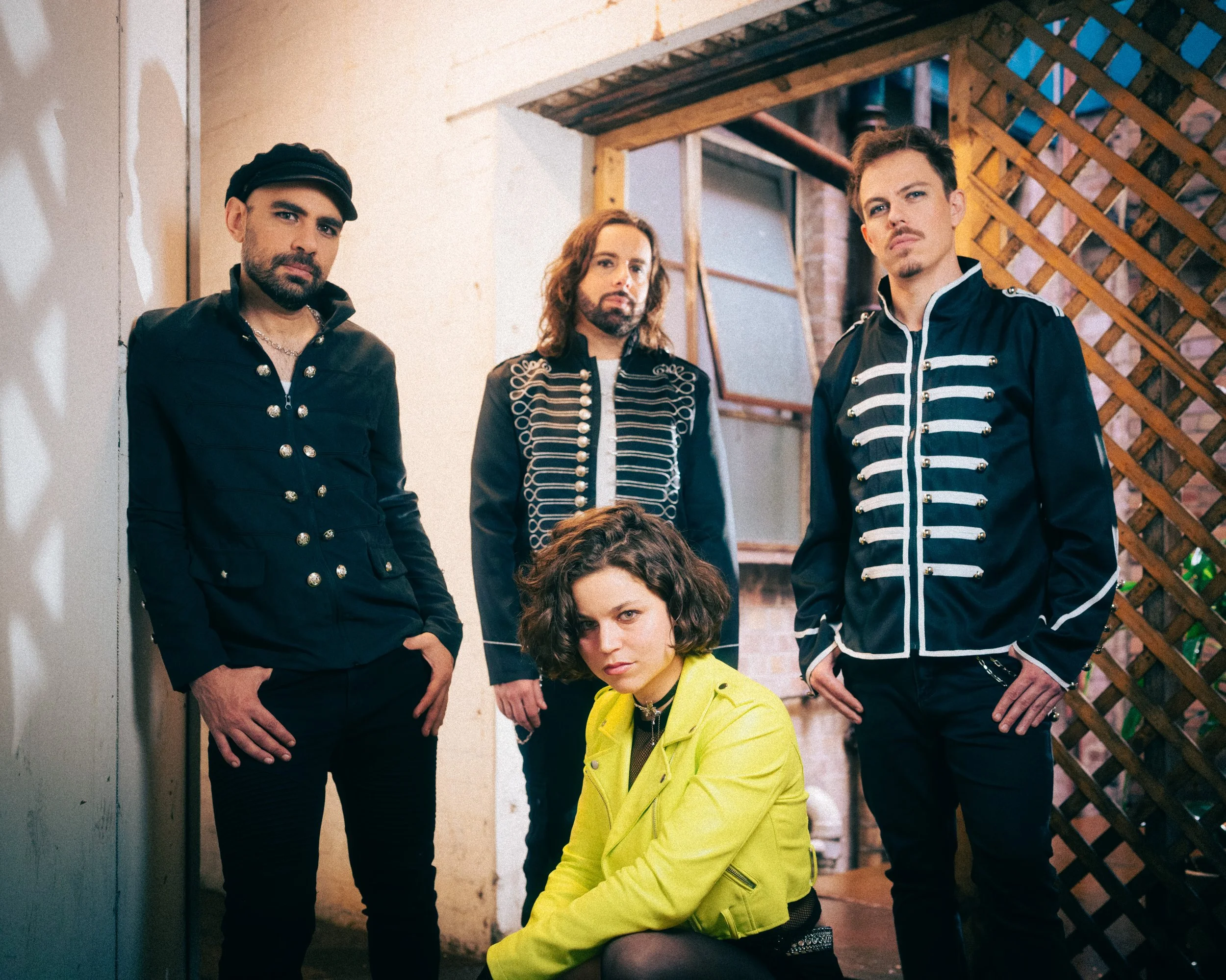 Five people, four men and one woman, pose together in an industrial-style setting. The woman is kneeling in front, wearing a bright yellow jacket. The men are standing behind her, dressed in black military-style jackets with silver embellishments. The background features exposed brick, wooden beams, and lattice fencing.