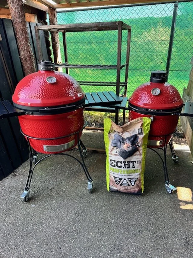 Two red Big Green Egg ceramic grills on a patio, with a bag of charcoal between them and a folding table in the background.