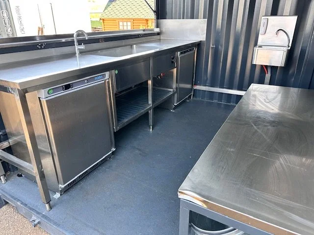Commercial stainless steel kitchen with sinks, under-counter refrigeration, and a table.
