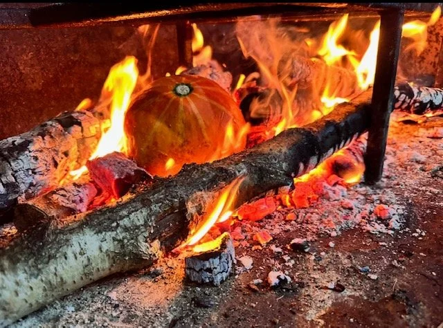 A pumpkin burning on an open fire with flames and charred logs around it.