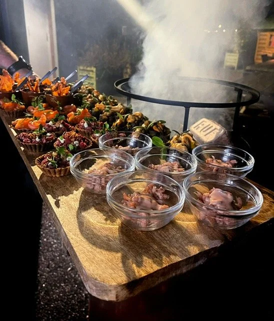 A barbecue grill with smoke in the background, a wooden serving board with assorted appetizers, including small cups of meat and topped with vegetables, some cups of raw meat, and other finger foods.