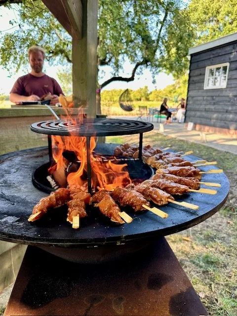 Close-up of a barbecue grill with skewers of meat cooking over open flames outdoors, with a man in a maroon shirt in the background and a wooden shed nearby.