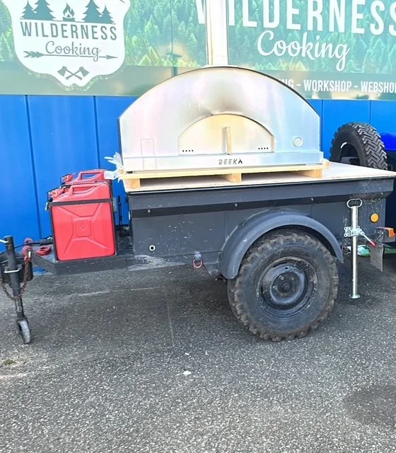 A portable outdoor pizza oven mounted on a trailer with a red fuel container attached, parked on a wet pavement near a blue wall with outdoor cooking signs.