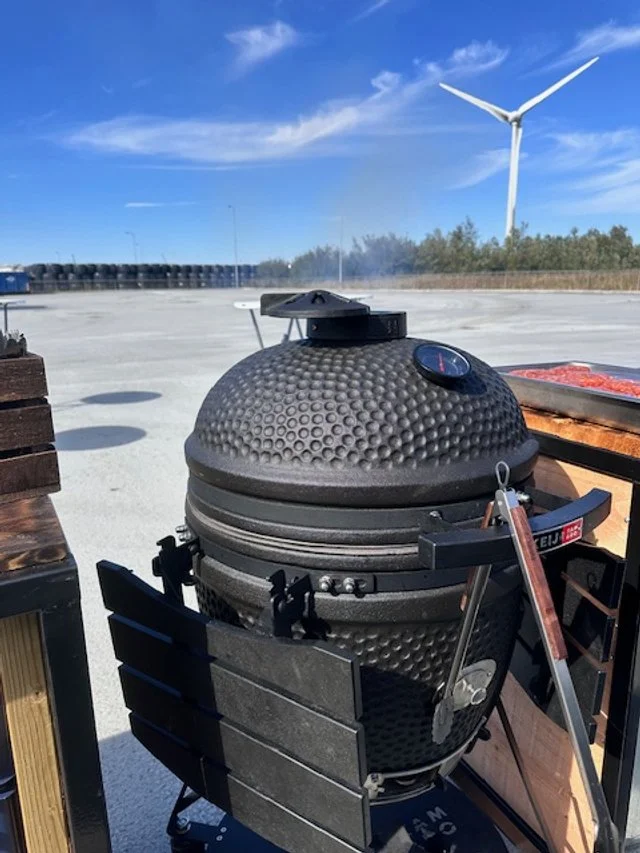 A black kamado-style grill with a dome shape and textured surface outdoors on a concrete surface, with wind turbines and a blue sky in the background.