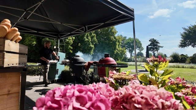 Outdoor cooking station with two smokers under a canopy, person preparing food, pink flowers in foreground, green trees and a grassy field in the background.