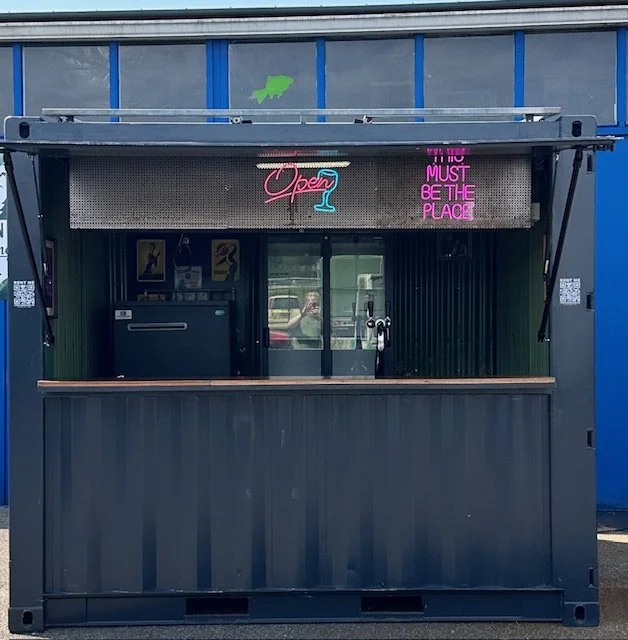 Empty outdoor bar booth with neon signs and a sliding glass window, blue and gray building facade in background.