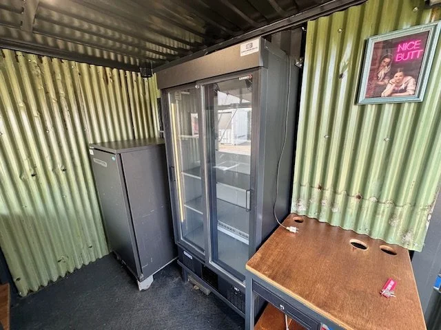 Empty vending cooler with glass doors next to a small black refrigerator, a wooden table with three cup holes and a pink lighter, and a framed poster on a green corrugated metal wall.