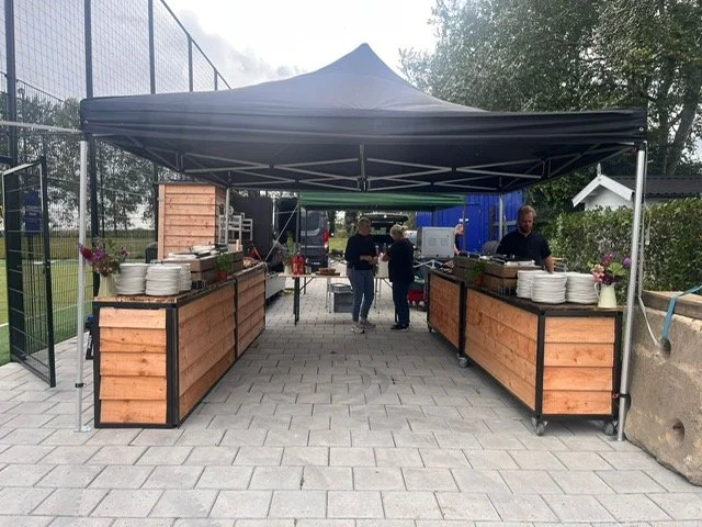 Outdoor catering setup with a black canopy, wooden counter serving food and drinks, and a few people gathered around in a park or garden area.