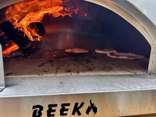 A pizza oven with flames inside, baking pizza. The oven is made of metal with the word 'BEEK' and a flame graphic on the front.