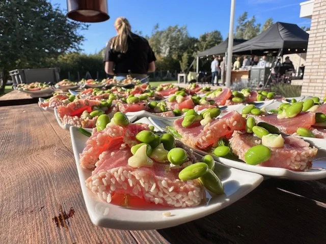 Close-up of rice salad with green edamame beans and diced tomatoes on white plates, set outdoors at a daytime gathering with people and tents in the background.