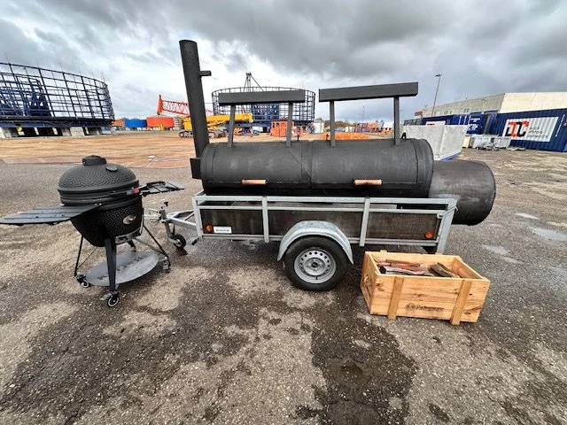 A black portable charcoal grill on a stand, a large black smoker attached to a trailer, and a wooden crate with grilling tools on an outdoor paved surface.