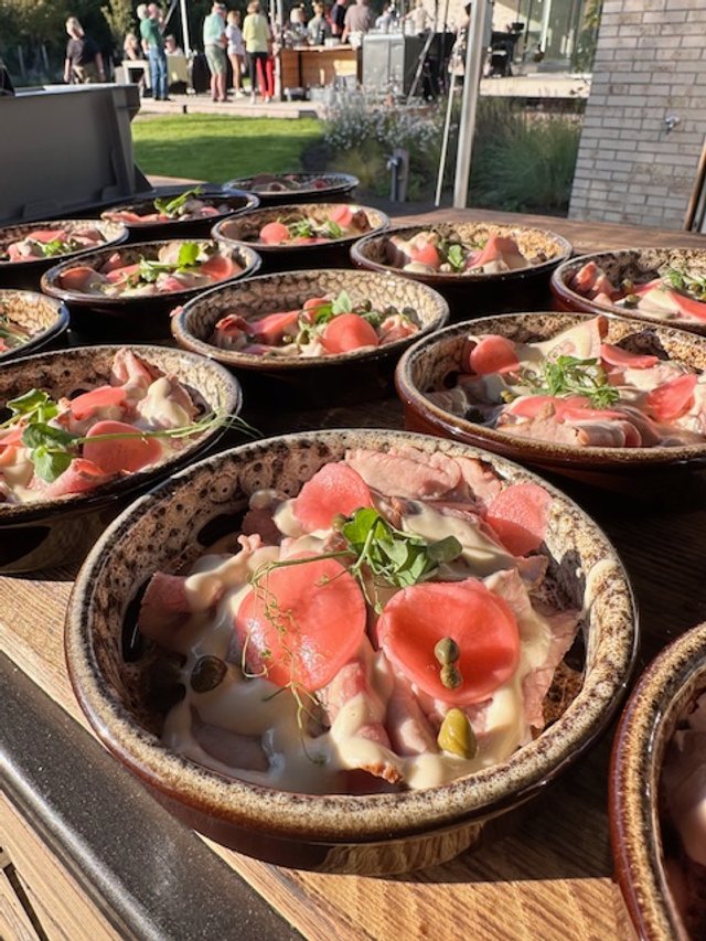 Multiple bowls of food with slices of meat, small green leaves, and pink or red garnish, set on a wooden table outdoors at a social gathering.