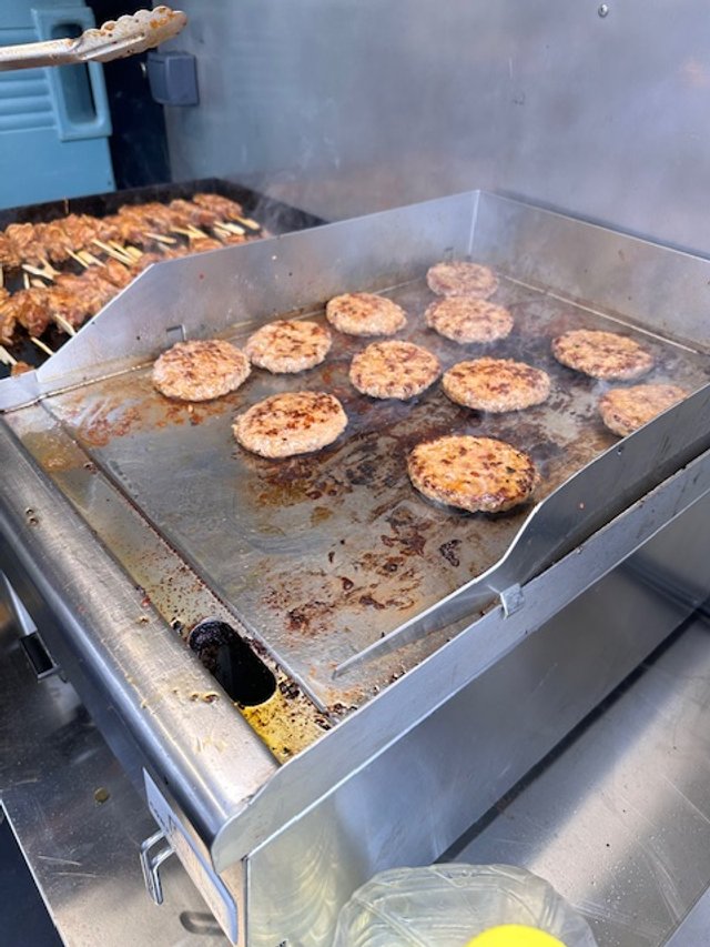 Ground meat patties cooking on a flat griddle with skewered meat in the background.