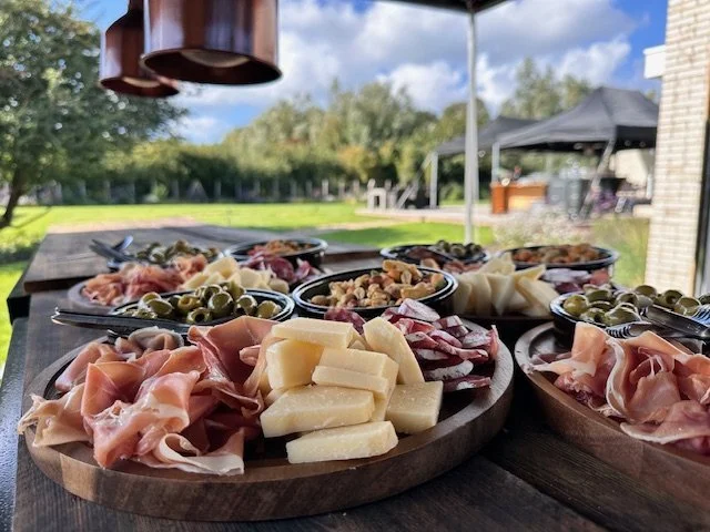 Charcuterie board with various cheeses, cured meats, and olives on a wooden table outdoors with a cloudy sky and tents in the background.
