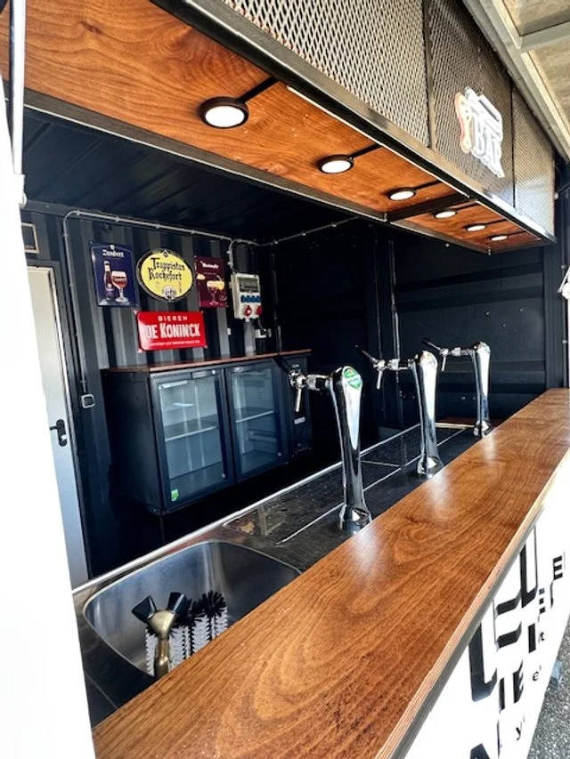 Indoor view of a bar with three beer taps, a sink with a brush, a mini fridge, and various beer tap signs on the wall.