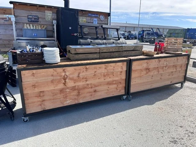 Outdoor food service station with a wooden counter, white plates, bowls, and condiment bottles, set up in a parking lot with trucks and a clear sky in the background.