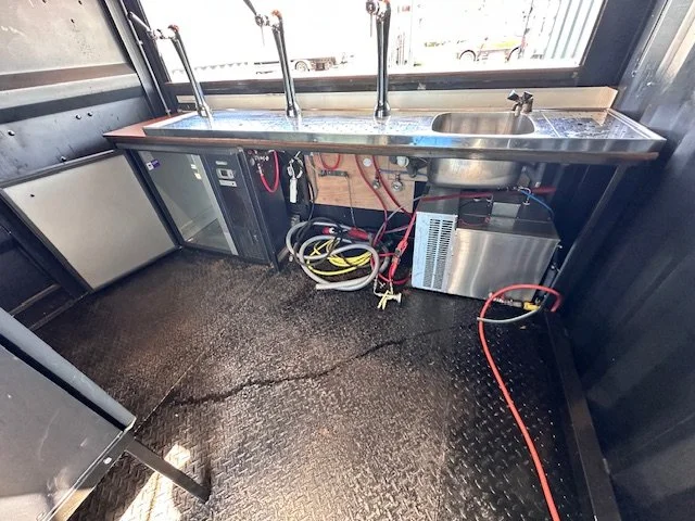 Commercial kitchen sink with two faucets, under-counter refrigerator, and electrical and plumbing connections in a mobile food truck or kitchen.