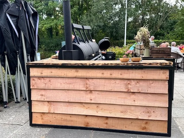 An outdoor wooden bar with a black grill in a park setting, surrounded by tables and trees.
