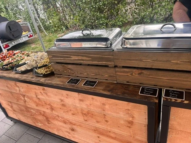 Buffet table with covered chafing dishes and trays of food, including pasta salads, with small black labels indicating food options, outdoors with greenery and trees in the background.