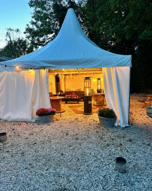 A white event tent with open flaps, illuminated from inside, set up outdoors on a gravel surface during evening. Inside, there are chairs, tables, and outdoor heaters, with potted plants at the entrance.