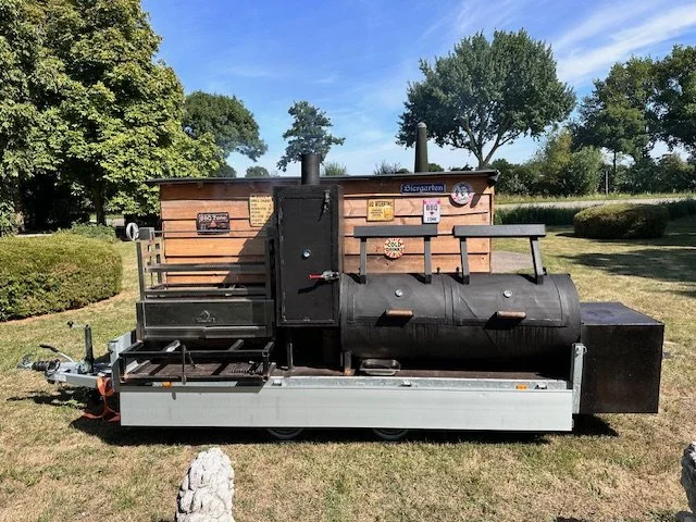 A mobile outdoor barbecue smoker with a large black cylindrical smoker, attached wooden panel, and signs, situated on a grassy area with trees and clear blue sky in the background.