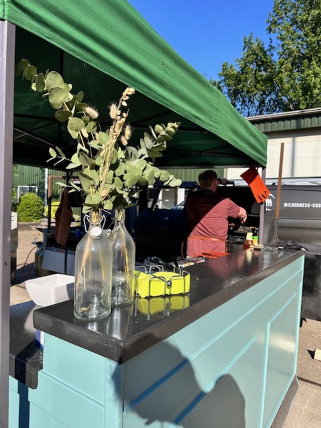 A food stand with a turquoise counter under a green canopy, decorated with glass bottles holding eucalyptus branches. There are two people behind the counter preparing food or drinks, with a bright, sunny day and clear blue sky in the background.