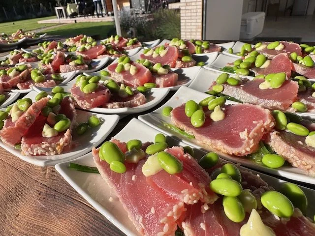 Several small white plates with slices of raw tuna and green edamame beans on top, arranged on a wooden table outside.