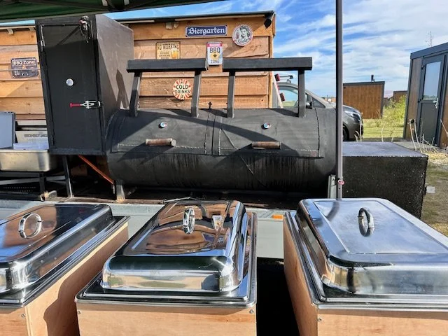 BBQ smoker grill in a food stand with three stainless steel chafing dishes in front, outdoor setting with signs and a blue sky.
