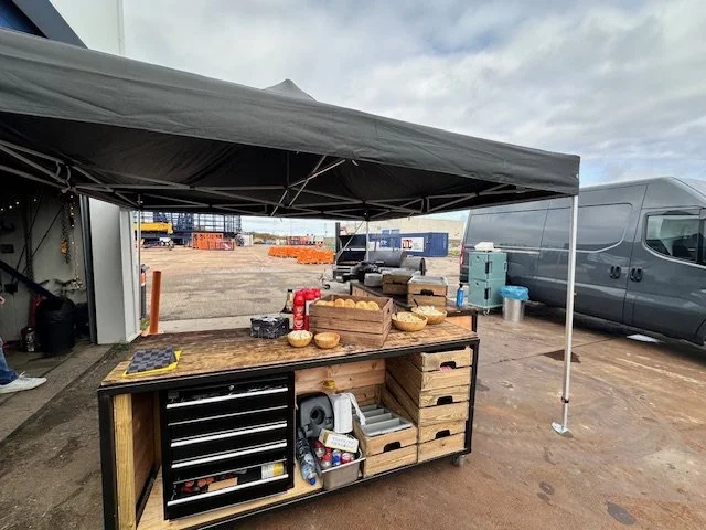 Outdoor workspace with a black workbench under a canopy, containing tools, wooden crates, bowls, and a fire extinguisher. In the background, there are construction materials, a large van, and a cloudy sky.