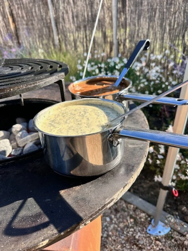 Two saucepans with dipping sauces on a black outdoor table next to a fire pit, with a garden and trees in the background.