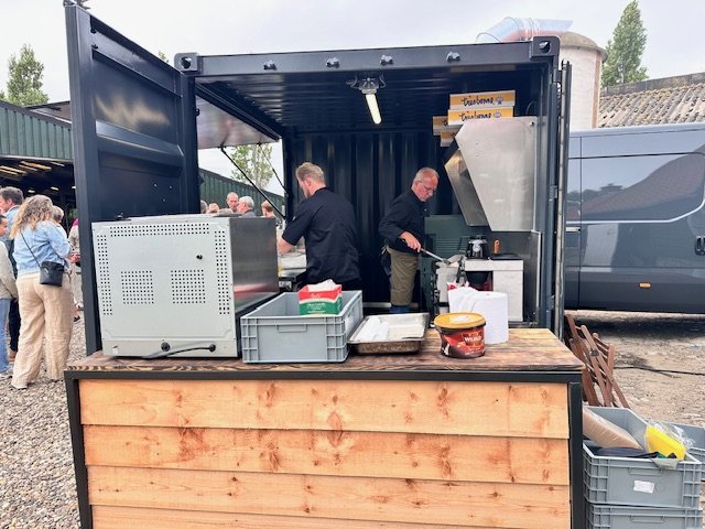 Two men working inside a food truck with a wooden counter, preparing food with external customers waiting in line.