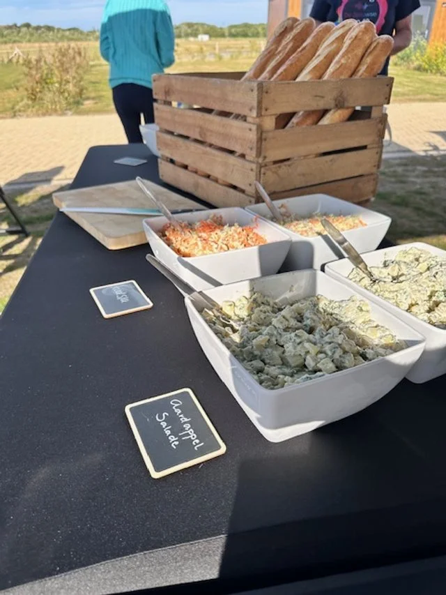 A buffet table with a basket of baguette bread and bowls of salad, including potato salad and crab salad, outdoors on a sunny day.
