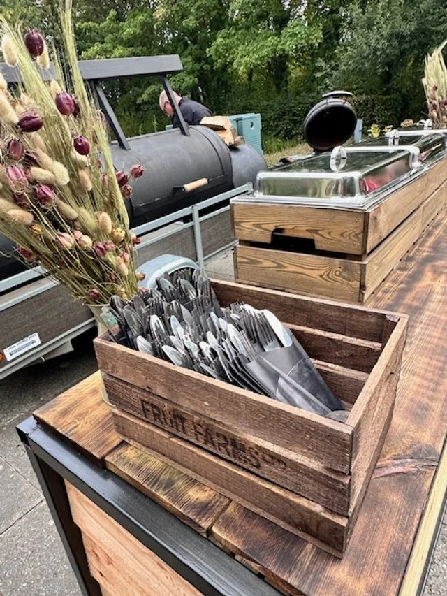 A rustic wooden box filled with metal grilling utensils, next to a bouquet of dried flowers, on a wooden table outdoors near a black smoker grill.