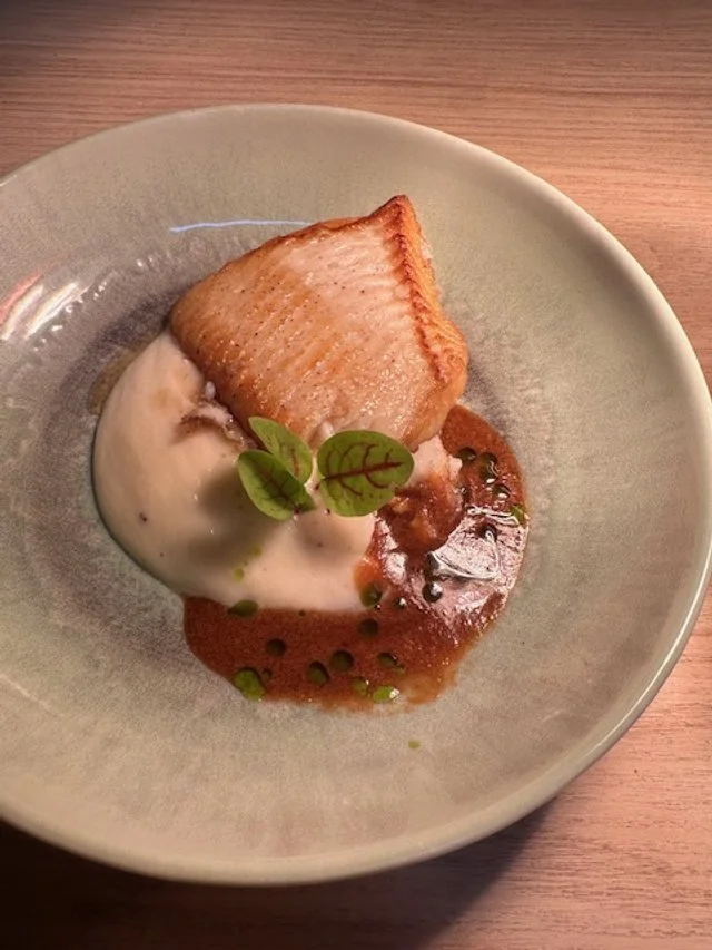 A plate with a piece of cooked fish, accompanied by a creamy sauce and garnished with small green leaves.