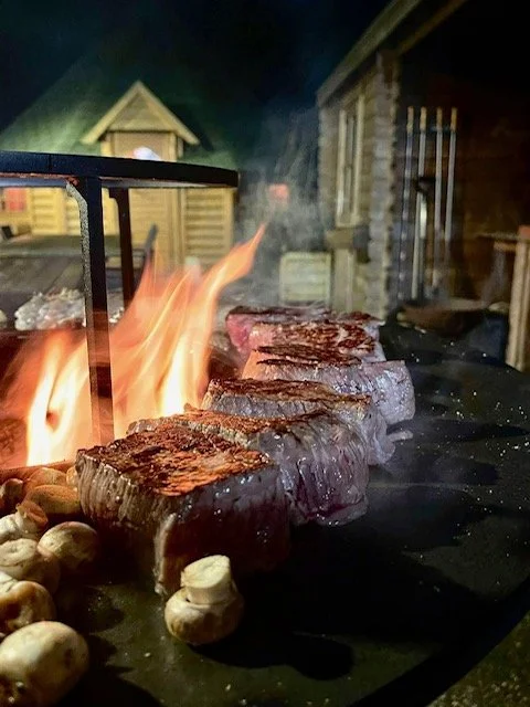Steaks cooking over an open flame on a grill, with mushrooms nearby, in an outdoor setting.
