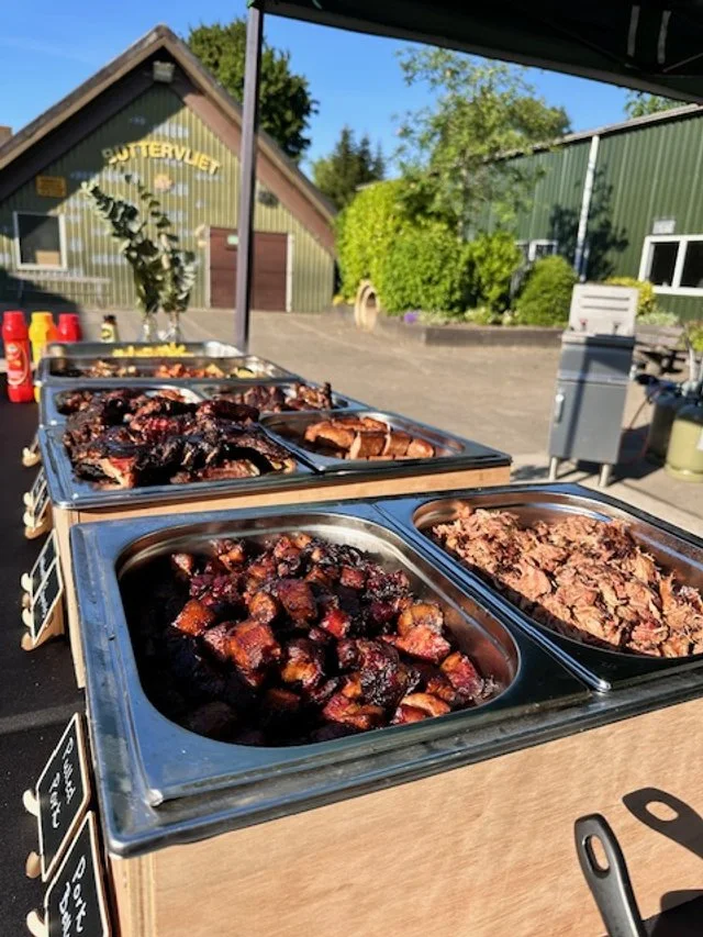 Buffet table with trays of grilled meats, including pork belly, brisket, and ribs, set outdoors near a green building and a barn with a sign that reads "Butterwett."