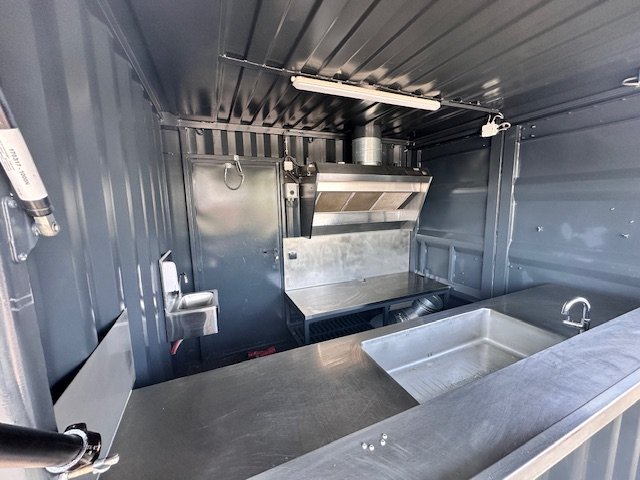 Interior of a stainless steel mobile kitchen with a sink, stove, and ventilation hood.