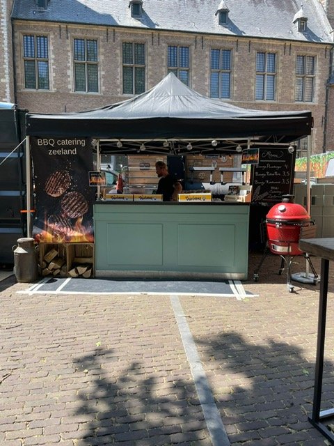 A small outdoor BBQ stand with a black canopy, a green front counter, a red grill, and a menu board, set up on a paved area in front of a large building.