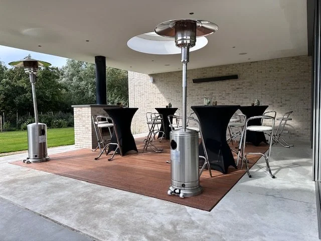 Outdoor patio with tall patio heaters, black cocktail tables, and metal chairs on a wooden rug, with a brick wall and green lawn in the background.