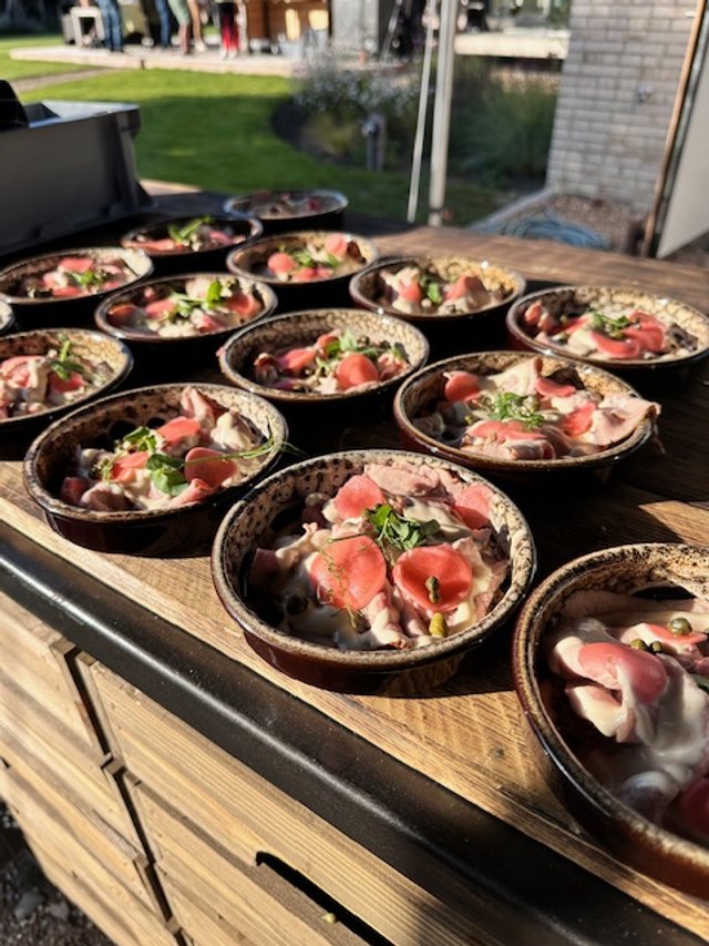 Multiple small bowls of food with sliced meat, cherry tomatoes, and herbs on a wooden table outdoors.
