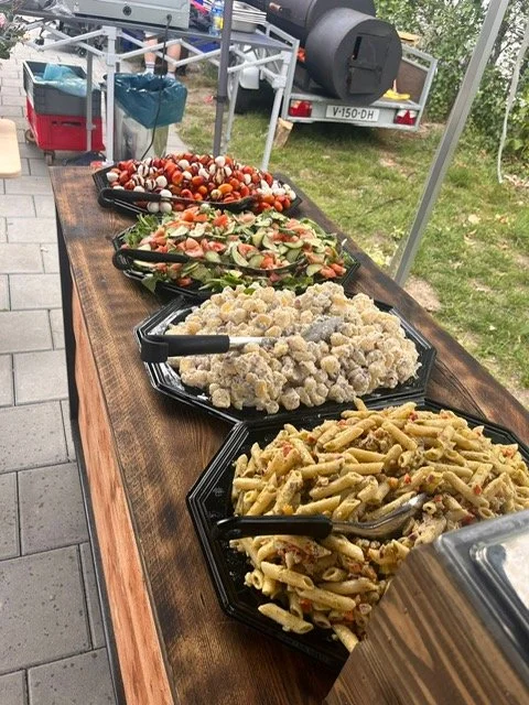 A long wooden table with five black trays filled with salads and pasta, outdoors at a gathering or event.