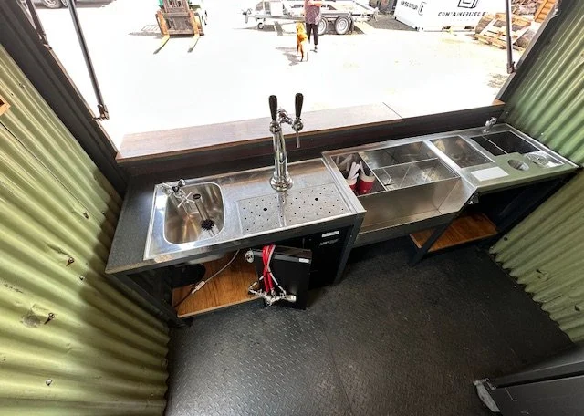 A small kitchenette area with a stainless steel sink, tap, and counter space, situated inside a metal shipping container with green corrugated walls. Outside, construction equipment and workers are visible.