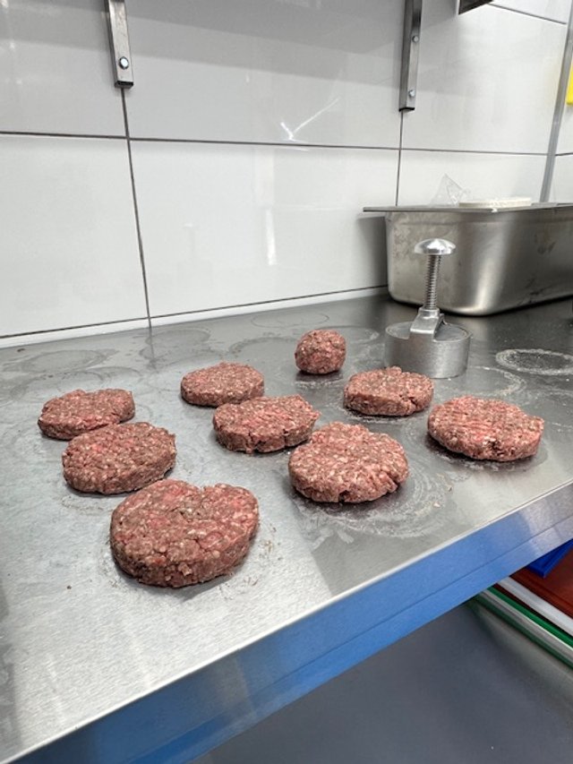 Ground beef patties on a stainless steel kitchen counter, with a metal weight and a large metal container nearby.